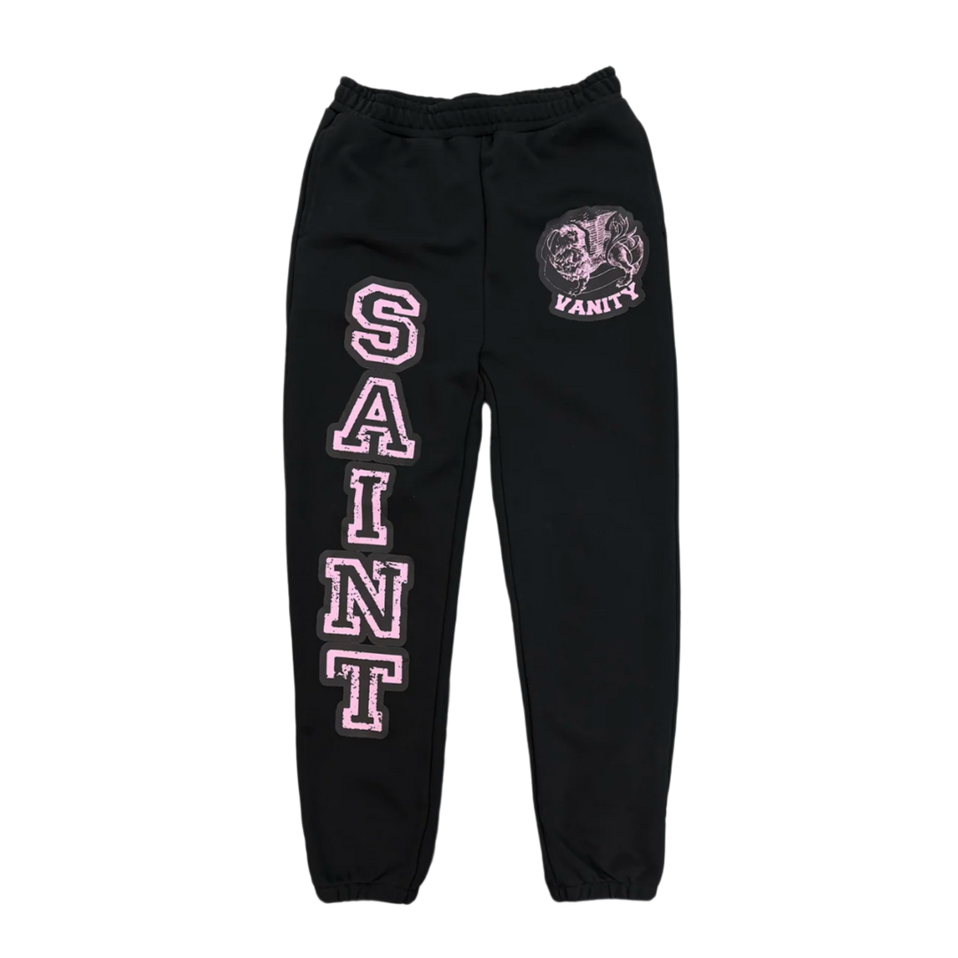 Front flat lay view of the Saint Vanity varsity sweatpants in heavyweight jet-black cotton fleece, showcasing a relaxed-taper lounge silhouette with gathered elastic ankle cuffs, a thick ruched elastic waistband, a large pink varsity block text graphic reading 'SAINT' running vertically down the right thigh, and a circular pink and white collegiate emblem graphic printed on the left hip.