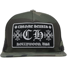 A straight front studio layout view of the Chrome Hearts trucker hat, showcasing a heavily structured woodland camouflage cotton twill front crown panel paired with breathable black mesh rear netting, a matching camouflage curved brim, and a polished sterling silver floral cross top button hardware accent, highlighted by a large embroidered front fabric patch featuring a bold white gothic 'CH' monogram logo framed by 'CHROME HEARTS' and 'HOLLYWOOD, USA' banners.