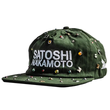 A three-quarter front angled studio layout view of the Satoshi Nakamoto x Sicko collaborative hat, showcasing a structured six-panel snapback cap profile crafted from a premium olive-green woodland camouflage cotton twill canvas, heavily embellished across the front crown and curved brim with an aggressive, scattered arrangement of polished silver metallic punk metal cone studs, and dominated by thick white embroidered block lettering on the center face reading 'SATOSHI NAKAMOTO' layered over Sicko's signat