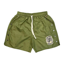 Front flat lay view of the Saint Vanity athletic shorts in structured matte olive green technical nylon fabric, showcasing a relaxed-fit mid-length silhouette, an elastic waistband with an adjustable long white contrast drawstring, functional metal snap-button closures running vertically along both outer side seams, and a circular grey and black embroidered collegiate coat-of-arms crest patch stitched near the lower left hem.