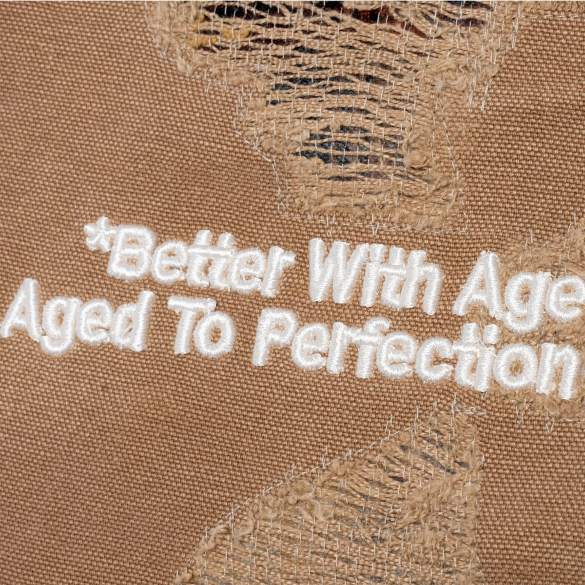 An extreme macro shot focusing on an intentionally distressed patch of the canvas fabric, showcasing frayed fiber layers overlaid with stylized white printed text that reads '*Better With Age Aged To Perfection'.