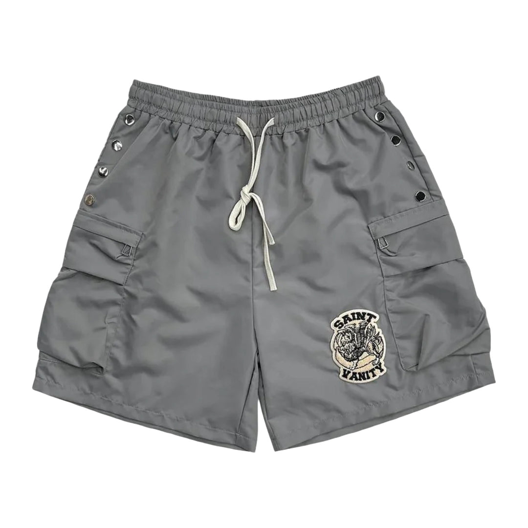 Front flat lay view of the Saint Vanity athletic shorts in structured matte slate grey technical nylon fabric, showcasing a relaxed-fit mid-length silhouette, an elastic waistband with an adjustable long white contrast drawstring, functional metal snap-button closures running vertically along both outer side seams, and a circular grey and black embroidered collegiate coat-of-arms crest patch stitched near the lower left hem.