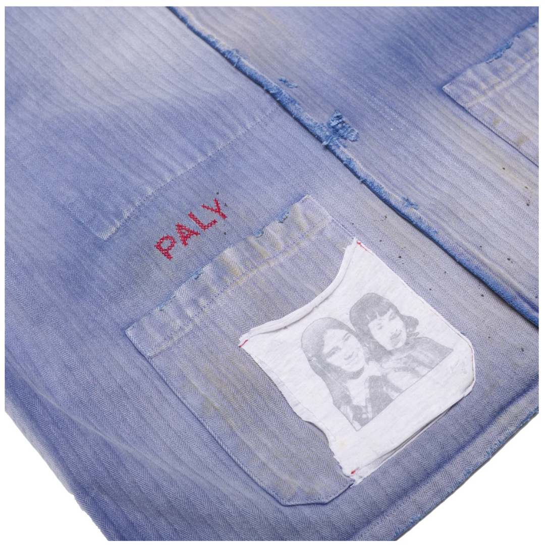 Macro detail of the lower front pocket on the Paly shirt, showing the frayed raw edges of the central seam, red 'PALY' chain-stitch embroidery, and a stitched white cotton patch featuring a vintage monochrome portrait.