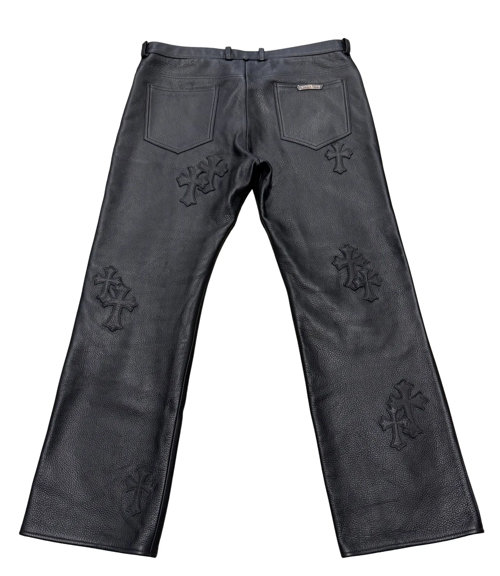 Back flat lay view of the black Chrome Hearts pebbled leather pants, showcasing the straight-leg silhouette with dual rear patch pockets, a silver scroll branding plaque on the right pocket, and a multi-layered arrangement of black leather cemetery cross patches stitched along the rear thighs and legs.