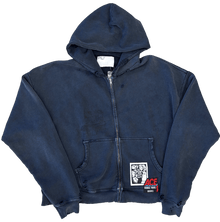 Front flat lay view of the Paly 'Theatre' full-zip hoodie in a vintage sun-faded navy blue cotton canvas, featuring an oversized boxy fit, dropped shoulders, a silver front zipper, split kangaroo pockets, and a graphic patch stitched onto the bottom right hem.