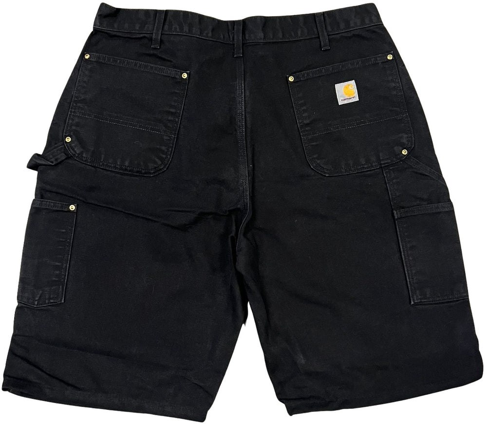 Back flat lay view of the black denim Carhartt carpenter shorts, showcasing two rear patch pockets with a woven Carhartt logo patch stitched onto the right pocket, alongside multi-layered tool utility pockets on the right thigh.
