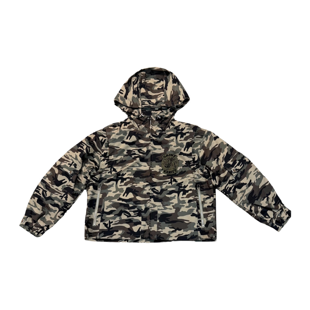 Front flat lay view of the Saint Vanity zip-up utility jacket in a textured woodland camouflage nylon fabric, showcasing an oversized boxy windbreaker silhouette with dropped shoulders, an integrated high-neck drawstring hood, a full-length black center zipper track, elasticated cuffs and lower hem, and multi-pocket technical detailing including traditional zip pockets paired with a small contrasting green embroidered patch applied to the left sleeve panel.