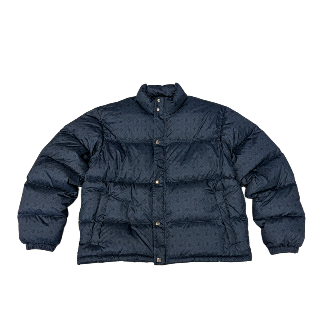 Front flat lay view of the Chrome Hearts down-filled puffer jacket in a deep navy blue technical satin shell, showcasing a voluminous insulated silhouette with wide horizontal insulation quilting segments, a plush stand-up protective collar, a concealed front zip closure placket with tonal button snaps, zippered hand pockets, and a subtle all-over tonal jacquard print of repeating miniature signature brand logos.