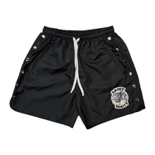 Front flat lay view of the Saint Vanity athletic shorts in structured matte black technical nylon fabric, showcasing a relaxed-fit mid-length silhouette, an elastic waistband with an adjustable long white contrast drawstring, functional metal snap-button closures running vertically along both outer side seams, and a circular grey and black embroidered collegiate coat-of-arms crest patch stitched near the lower left hem.