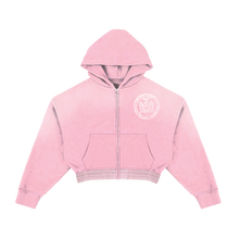 Front flat lay view of the Saint Vanity women's collegiate full-zip hoodie in a soft pastel pink cotton fleece, showcasing a cropped boxy silhouette with dropped shoulders, long relaxed sleeves with thick ribbed cuffs, a silver metal front zipper track, split kangaroo pouch pockets, a spacious slouchy hood, and a circular white collegiate emblem graphic printed subtly on the left chest.