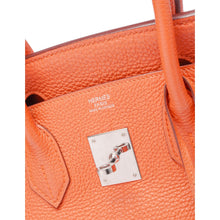 A view looking inside the open interior of the orange leather handbag, highlighting the matching orange leather lining and a slip pocket stamped with the silver foil metallic Hermès logo.