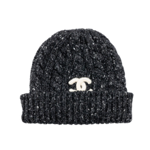 Front studio view of the Chanel beanie, showcasing a premium heavyweight cable-knit cashmere composition executed in a textured black and ecru cream mottled melange yarn pattern, featuring a thick rib-knit fold-over cuff design, a structured watch-cap skull silhouette, and a prominent stark white contrast interlocking 'CC' signature logo patch embroidered centered on the front cuff.