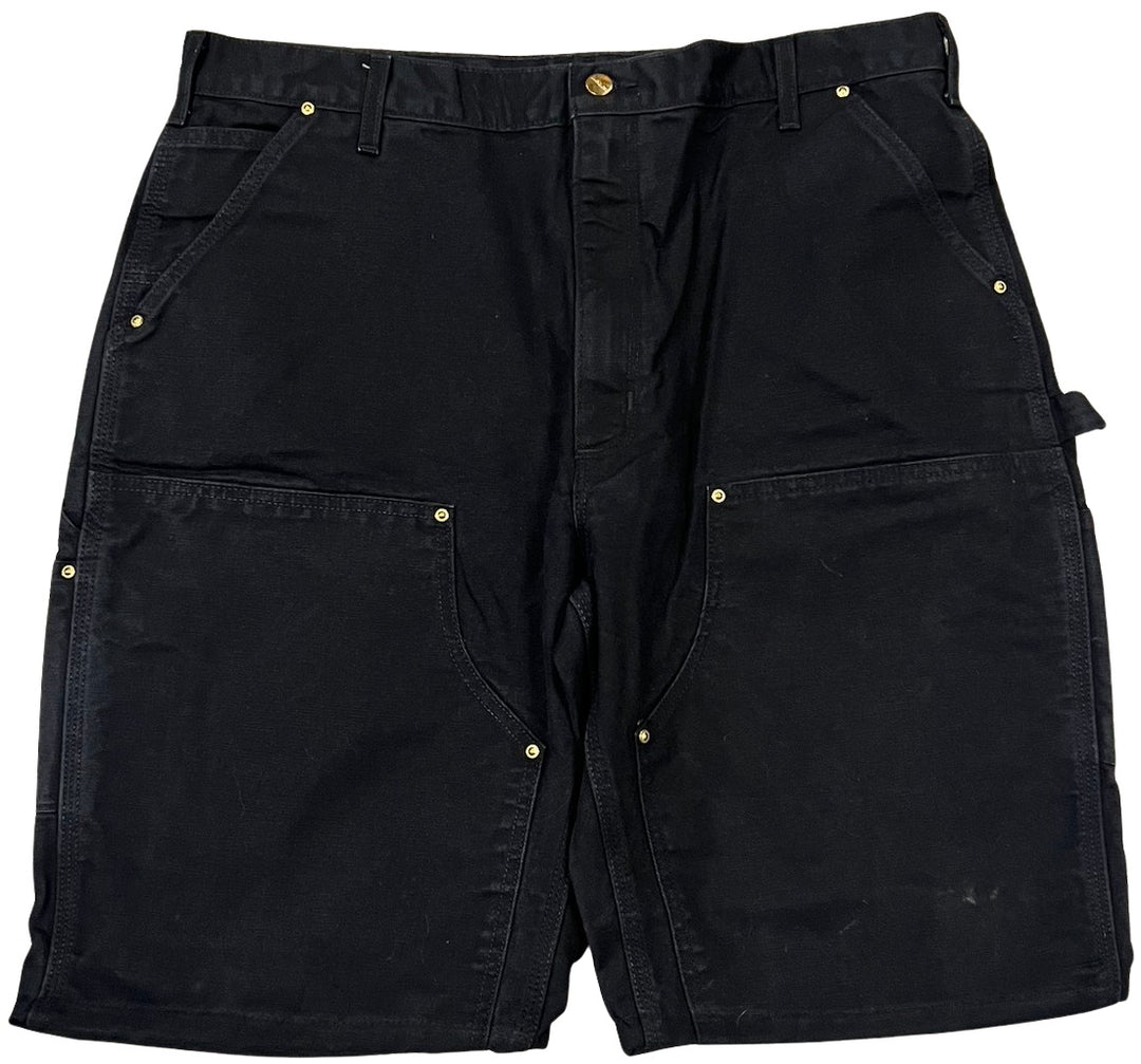 Front flat lay view of the Carhartt carpenter shorts in a heavy black washed denim, featuring reinforced double-knee front panels with metallic rivets, deep side slip pockets, and a classic hammer loop utility detail on the left leg.