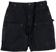 Front flat lay view of the Carhartt carpenter shorts in a heavy black washed denim, featuring reinforced double-knee front panels with metallic rivets, deep side slip pockets, and a classic hammer loop utility detail on the left leg.