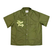 Front flat lay view of the Saint Vanity button-up shirt in smooth matte olive green technical nylon fabric, showcasing a boxy short-sleeve silhouette, a structured pointed camp collar, a full-length olive button front closure placket, a square flat hemline, and elegant light yellow cursive script branding reading 'Saint Vanity' embroidered directly over the right chest.
