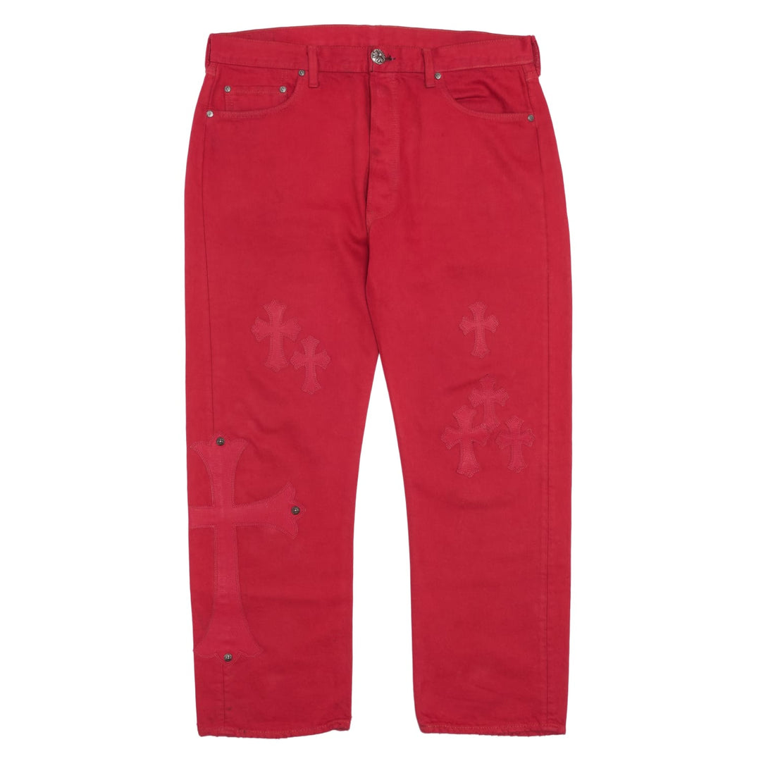 Front flat lay view of the Chrome Hearts tailored denim jeans in a striking, deep cherry-red garment dye, showcasing a straight-leg streetwear silhouette decorated down both legs with an asymmetrical arrangement of hand-stitched tonal red leather cemetery cross patches.
