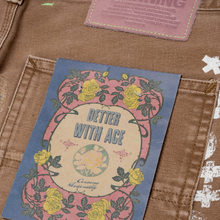 A detailed close-up shot of the rear right patch pocket area, emphasizing the unique color combination of a stitched circular vintage-style graphic badge nestled near a smaller pastel pink square branding hit.