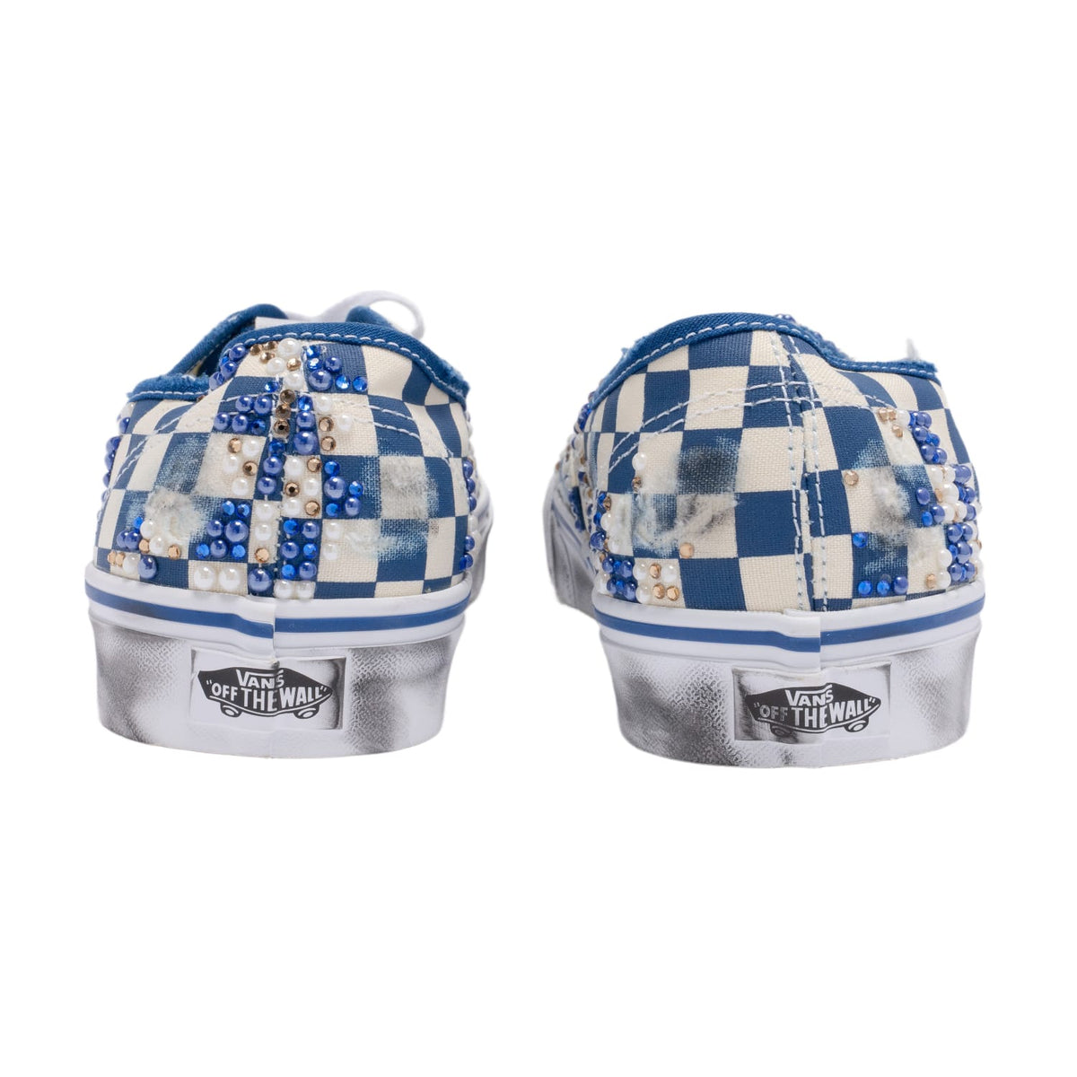 A straight-on rear view of the pair of sneakers placed side-by-side, showcasing the vertical heel accent strips in distressed blue checkerboard canvas and the classic red rubber 'Vans Off The Wall' license plate heel logo plaques applied to the base of the white vulcanized midsoles.