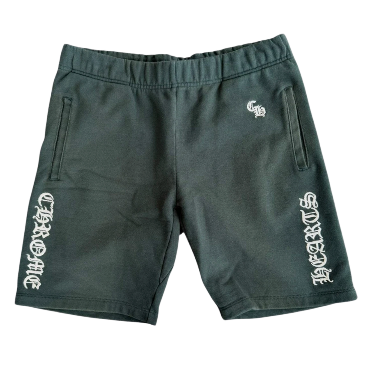 Front flat lay view of the Chrome Hearts 'Slo Ride' shorts in dark forest green cotton fabric, featuring a gathered elastic waistband, a small white embroidered gothic monogram on the upper left hip, and matching vertical white gothic 'CHROME HEARTS' script framing both lower leg openings.