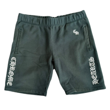 Front flat lay view of the Chrome Hearts 'Slo Ride' shorts in dark forest green cotton fabric, featuring a gathered elastic waistband, a small white embroidered gothic monogram on the upper left hip, and matching vertical white gothic 'CHROME HEARTS' script framing both lower leg openings.