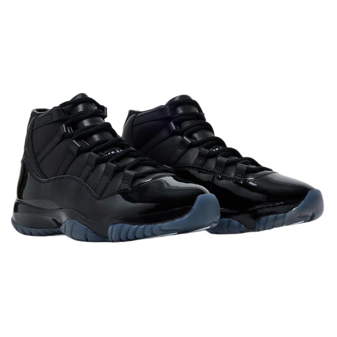 A pair of monochrome black Comme des Garçons x Air Jordan 11 sneakers displayed at a three-quarter angle, highlighting the contrast between the matte black premium leather upper, the glossy patent leather mudguard, and a semi-translucent smoky blue rubber outsole.
