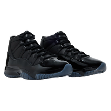 A pair of monochrome black Comme des Garçons x Air Jordan 11 sneakers displayed at a three-quarter angle, highlighting the contrast between the matte black premium leather upper, the glossy patent leather mudguard, and a semi-translucent smoky blue rubber outsole.