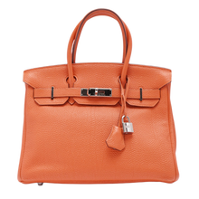 Front view of the Hermès Birkin 30 handbag in iconic textured orange grained leather, featuring dual rolled leather top handles, a closed front flap, a leather clochette with keys, and a silver palladium turn-lock closure.