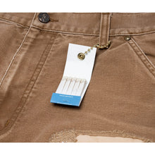 A detailed close-up view of the outer side seam, highlighting a functional white paper retail hangtag attached near the pocket, the thick canvas texture, and the clean edge geometry of the utility tool pocket construction.