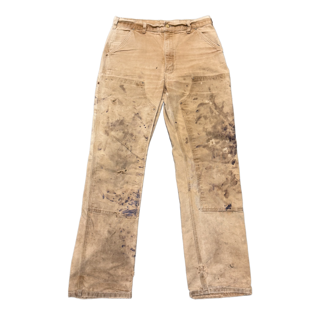 Front flat lay view of vintage Carhartt double-knee work pants in heavily distressed tan duck canvas, showcasing a rugged straight-leg utility silhouette, a classic multi-pocket layout, reinforced protective double-layer canvas panels extending across the thighs and knees, a traditional button-and-zip fly waistband, and authentic heavy grease stains, fading, and hand-applied white paint splatters throughout the canvas material texture.